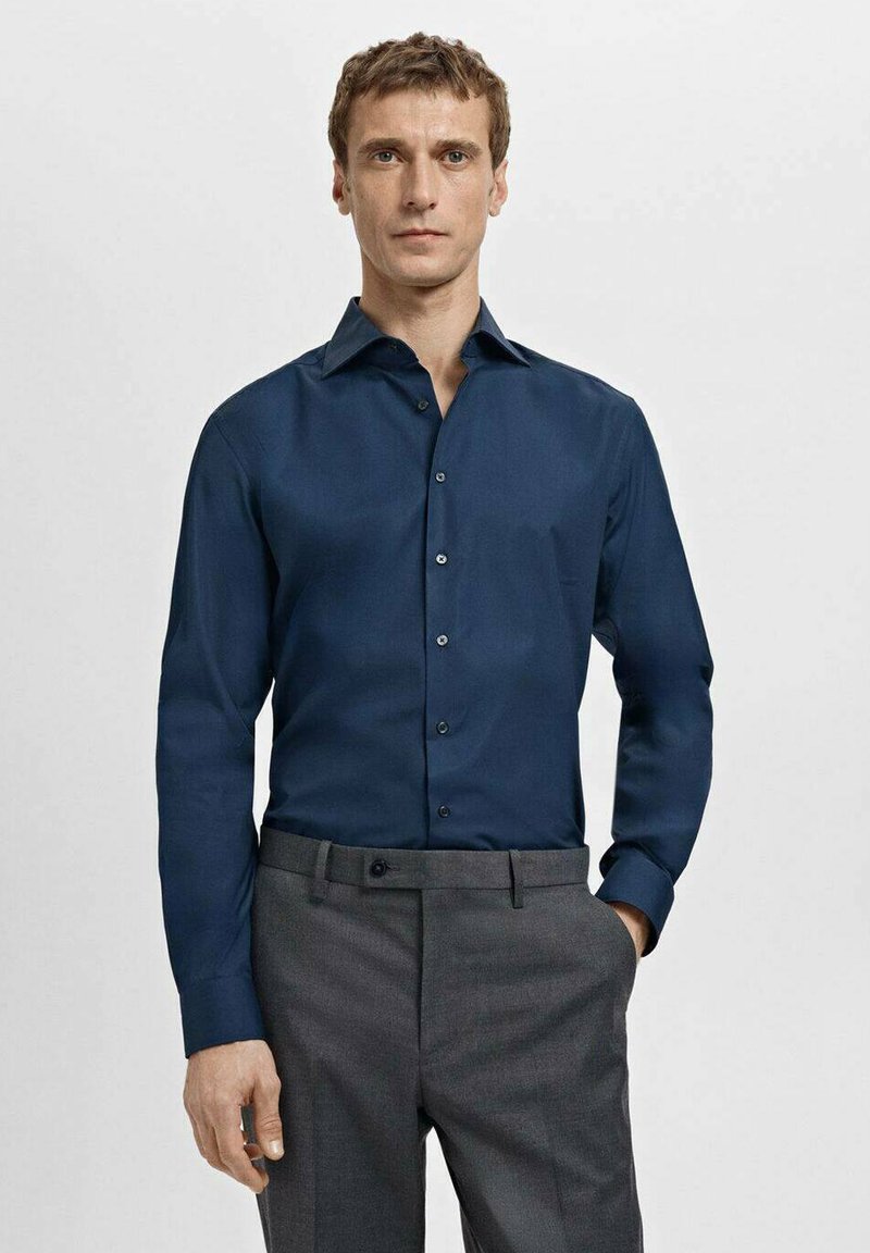 Granatowa koszula z długim rękawem, kołnierzem typu spread, plisą na guziki i gładką tkaniną, zestawiona z szarymi, dopasowanymi spodniami.