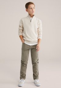 Šviesiai smėlio spalvos megztinis su ketvirčio užtrauktuku, kreminės spalvos vidinis sluoksnis, alyvuogių žalumos kelnės ir šviesiai mėlyni sportbačiai. Lygios tekstūros su ribuotu rankogaliu ir juosmeniu.