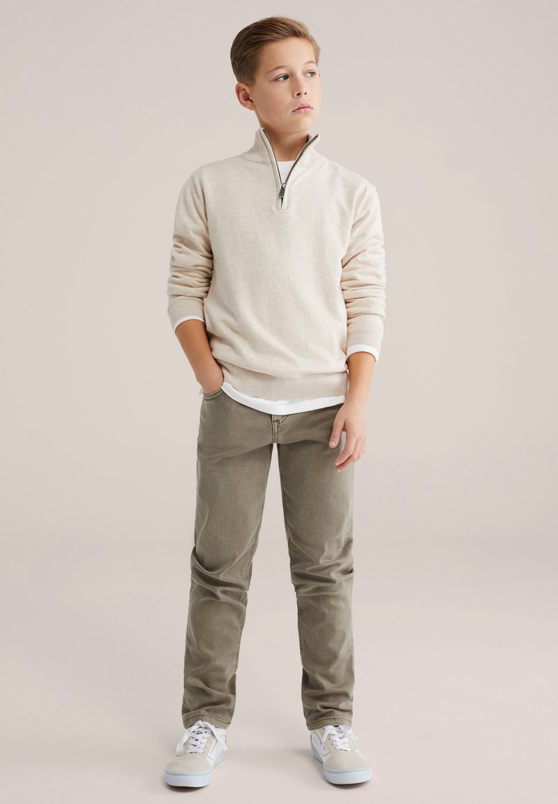 Šviesiai smėlio spalvos megztinis su ketvirčio užtrauktuku, kreminės spalvos vidinis sluoksnis, alyvuogių žalumos kelnės ir šviesiai mėlyni sportbačiai. Lygios tekstūros su ribuotu rankogaliu ir juosmeniu.