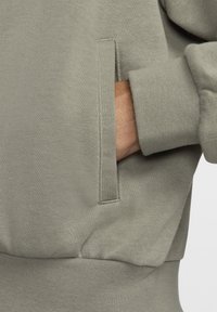 Felpa verde oliva con tessuto strutturato, polsini a coste e una tasca laterale con cucitura verticale all'apertura. Superficie del materiale liscia.