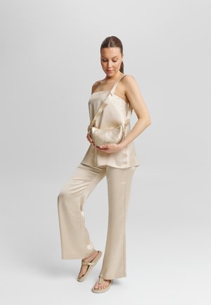 Satin-beige Top mit Spaghettiträgerschnitten und weiten Hosen, kombiniert mit einer metallic Minitasche. Das Model posiert lässig und trägt flache Sandalen.