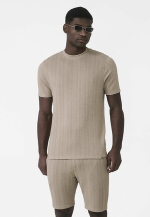 Beżowy dzianinowy top z krótkim rękawem o pionowym, fakturowanym wzorze, w zestawie z pasującymi szortami. Modelka nosi ciemne okulary przeciwsłoneczne.