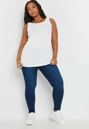 Frau steht und trägt ein weißes ärmelloses Oberteil, dunkelblaue Skinny-Jeans und weiße Turnschuhe vor einem schlichten weißen Hintergrund.