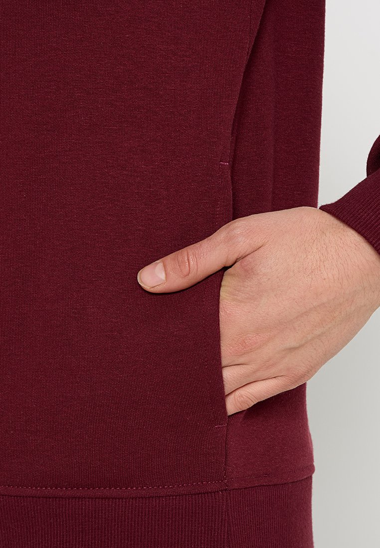 Bordeauxfarbener Pullover mit einer Seitentasche, gerippten Bündchen und einer weichen Textur. Nahaufnahme einer Hand, die in der Tasche ruht.