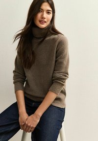 Suéter de cuello alto en marrón claro, hecho de una tela suave y texturizada, con puños y dobladillo acanalados, combinado con jeans azul oscuro.