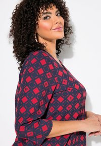 Top a righe blu navy e rosso con scollatura a V e maniche tre quarti. Presenta forme geometriche e dettagli testuali su tutta la stoffa.