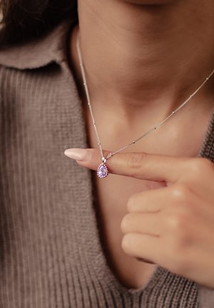 Silberne Halskette mit einem lila Tropfenanhänger, der fein ausgearbeitet ist. Hand positioniert nahe dem Anhänger, um das Design zu zeigen.