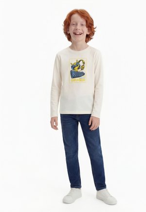 Osemdesetletni deček z rdečimi lasmi, ki se smeji, oblečen v belo majico z dolgimi rokavi z grafiko iger, modre kavbojke in bele superge, stoji pred belim ozadjem.