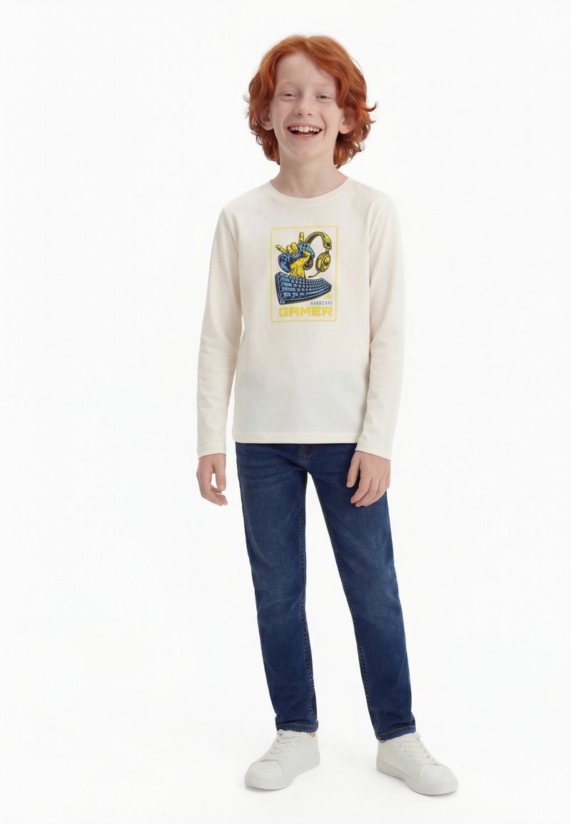 Κόκκινο-μαλλιαρό αγόρι που χαμογελά, φορώντας ένα λευκό μακρυμάνικο μπλουζάκι με γραφική απεικόνιση παιχνιδιών, μπλε τζιν και λευκά αθλητικά παπούτσια, στέκεται μπροστά σε λευκό φόντο.