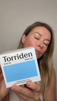 Donna con capelli biondi lunghi che tiene una scatola del prodotto per la cura della pelle Torriden Multi Pad, occhi chiusi, su sfondo chiaro e uniforme.