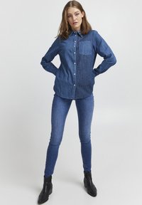 Camicia di denim con design button-down, due tonalità di blu, maniche lunghe, taschino sul petto, indossata con jeans skinny e stivali alla caviglia.