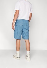 Man i blå cargoshorts, vita knästrumpor, vita höga sneakers och en enkel vit kortärmad t-shirt, stående mot en vit bakgrund.