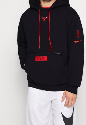 Man draagt een zwarte Chicago Bulls hoodie met rode trekkoorden en logo, en witte Nike shorts met zwarte swoosh, staand tegen een witte achtergrond.