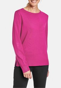 Uśmiechnięta osoba w jasnoróżowym swetrze z długim rękawem, z bocznymi rozcięciami i czarnych spodniach, stojąca na tle białym.