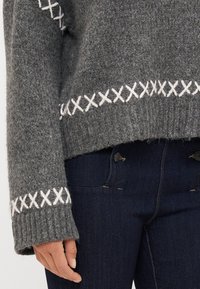 Šedý pletený svetr s bílým vyšíváním na lemování a manžetách, s ribbed texturou a volným střihem, v kombinaci s modrými džíny.