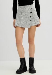 Minifalda de tweed gris con un patrón texturizado, que presenta cinco botones negros y un dobladillo asimétrico. Combinada con botas negras de cordones.