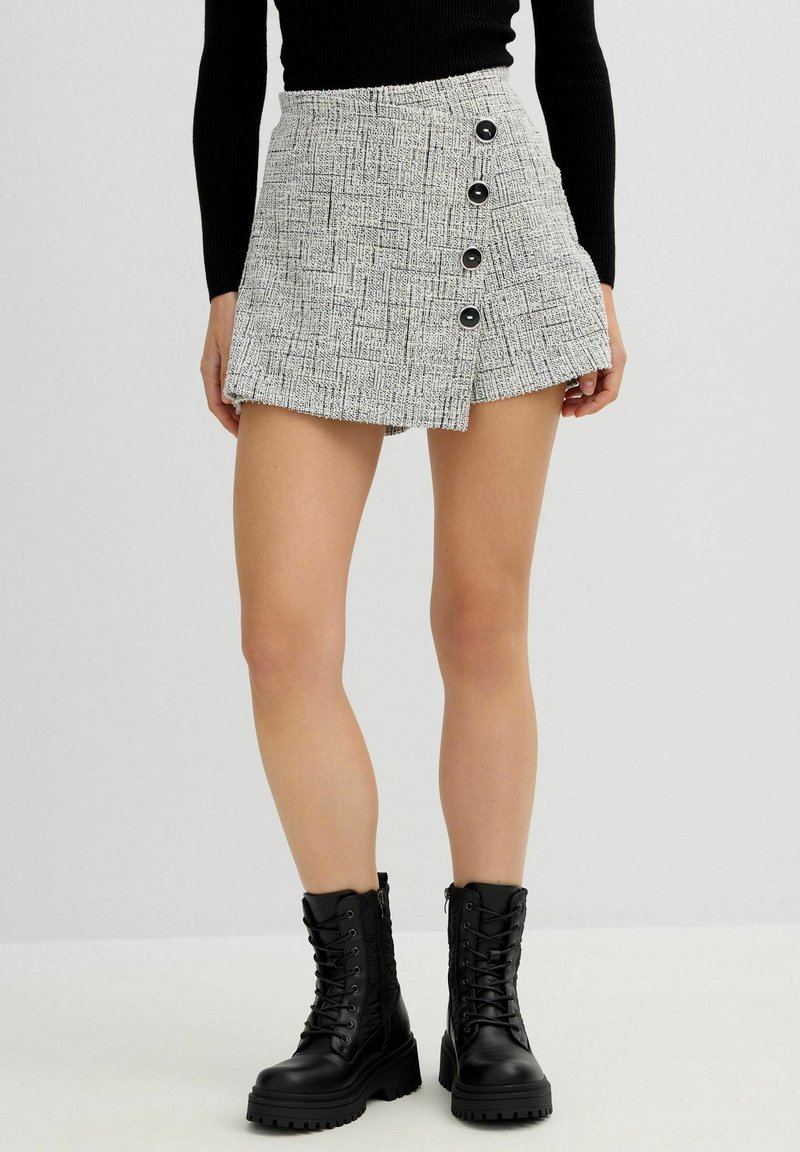 Minifalda de tweed gris con un patrón texturizado, que presenta cinco botones negros y un dobladillo asimétrico. Combinada con botas negras de cordones.