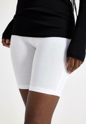 Kvinde iført hvide tætsiddende knælange shorts og en sort langærmet top, står med den ene hånd afslappet ved siden.