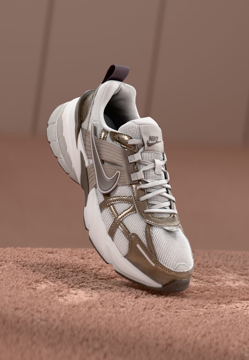 Zapatillas Nike con una parte superior de malla en blanco, con acentos metálicos en bronce. Diseño de suela gruesa y detalles en cordones grises.