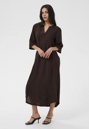 Femme aux longs cheveux foncés debout, portant une robe midi ample marron foncé et des sandales à talons noires à brides, sur un fond clair uni.