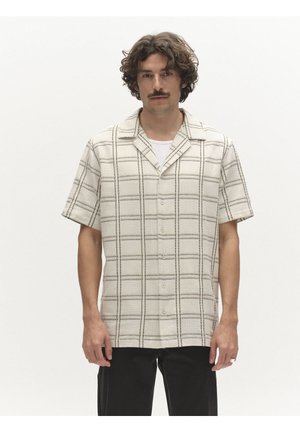 Göndör hajú és bajszos férfi, aki bézs színű, fekete rácsmintás rövid ujjú gombos ingben és fekete nadrágban áll, fehér háttér előtt.