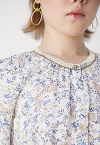 Kvinne som har på seg gulldobbel hoop øredobber og en hvit bluse med blå og rosa blomster- og paisleymønster, med små knapper og en perlekant.