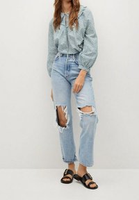 Helles blaues, gemustertes Blusenshirt mit Volant-Ausschnitt, kombiniert mit mittelblauen, ausgebeulten Boyfriend-Jeans. Schwarze flache Sandalen mit goldenen Beschlägen.