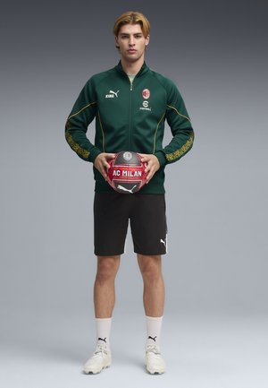 Groen-gele ritssluiting jas met AC Milan-logo, zwarte korte broek, witte sokken en lichte schoenen, houdend een rood-witte voetbal vast.