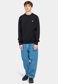 Ung man som står iklädd en svart sweatshirt, lösa blå jeans och svarta skor mot en ljusgrå, enhetlig bakgrund.