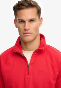 Sudadera roja de media cremallera hecha de una tela texturizada, con costuras en contraste y un logo en el pecho. Forro interior suave visible.