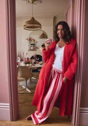 Rote, strukturierte, lange Robe mit großen Taschen, kombiniert mit rosa-weiß gestreiften Schlafanzughosen. Hält eine rosa Tasse und steht in einer Tür.