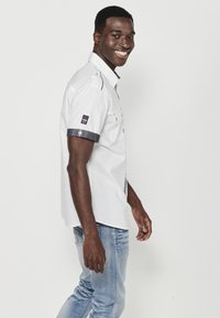 Camisa blanca de manga corta con cuello tipo polo, dos bolsillos en el pecho y detalles de mezclilla en las mangas. Se lleva con jeans de color azul claro.