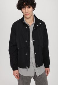 Black lightweight jacket with a zip front, snap fasteners, and side pockets, paired with a black and white striped shirt underneath.