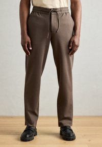 Brune, tapered bukser lavet af blødt stof med elastisk talje og snor. Har sidelommer og en glat tekstur.
