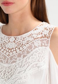 Blouse blanche avec un col en dentelle florale complexe et un tissu transparent. Présente une délicate broderie et un drapé doux. Pas de fermetures visibles.