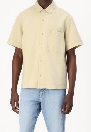 Camisa de botones de manga corta en suave tejido beige, con un cuello clásico y un bolsillo en el pecho con un discreto detalle del logo.