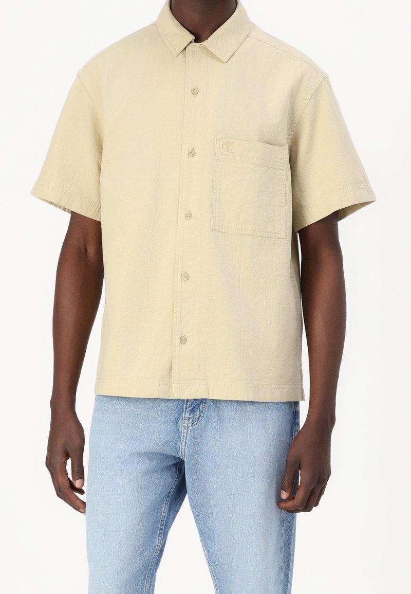 Korte mouwen blouse in zacht beige stof, met een klassieke kraag en een borstzak met subtiel logo detail.