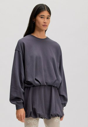 Femme aux longs cheveux foncés portant un sweat-shirt gris foncé oversize et des leggings en dentelle florale beige clair, debout devant un fond clair uni.
