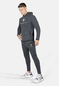 Mørk grå hettegenser med frontlomme og logo. Matchende slim-fit grå joggebukser. Modell som har på seg hvite og grå treningssko.