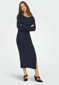 Abito a costine blu navy con maniche lunghe e una fessura laterale, caratterizzato da un ampio scollo. Abbinato a mule con tacco neri.