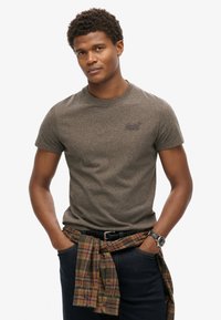Kortærmet, gråbrun t-shirt med rund halsudskæring og broderet logo; ternet skjorte bundet omkring taljen over mørke jeans.