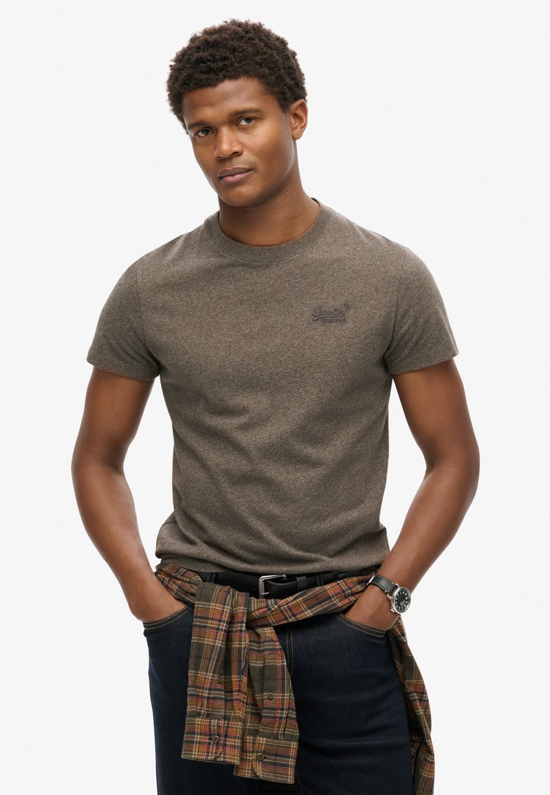 Korte mouwen, grijsbruine t-shirt met een ronde hals en geborduurd logo; geruite blouse om de taille geknoopt over donkere jeans.