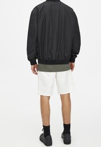 Veste bomber noire avec poignets élastiques, portée sur une chemise verte et un short blanc. Baskets noires et chaussettes foncées complètent la tenue.