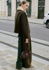 Abrigo de lana marrón con un cuello de piel de oveja crema, combinado con pantalones a juego. Lleva un bolso de cuero marrón oscuro con detalles de herrajes dorados.