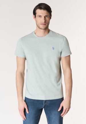 Uomo che indossa una maglietta a maniche corte grigio chiaro con un piccolo logo blu sul petto a sinistra e jeans blu, in piedi contro uno sfondo semplice.