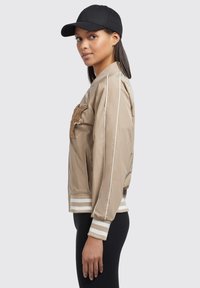 Beige bomberjack met geborduurd patch, contrasterende witte strepen op de manchetten, ritssluiting en zijzakken. Draag met een zwarte baseballcap.