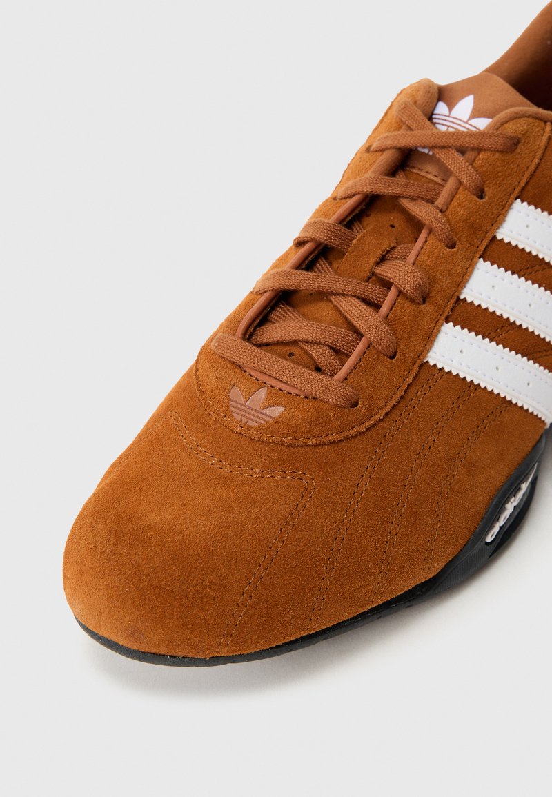 Bruine suède sneaker met een wit ontwerp van drie strepen, met een afgeronde neus en een gestructureerd bovenwerk. Zwarte rubberen zool met merkdetails.