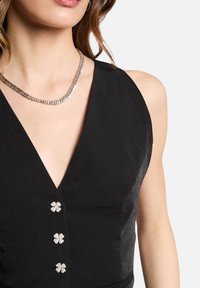 Haut sans manches noir avec un décolleté en V profond, présentant trois boutons en métal argenté en forme de fleur et un tissu lisse et texturé.