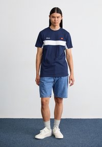 Marineblå bomulds T-shirt med hvid vandret stribe og logo, parret med denimshorts. Modellen har hvide sneakers og lysegrå sokker på.