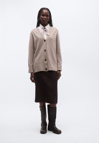 Beež värvusega kootud kampsun, millel on V-kaelus ja pruunid nupud, kantud valge särgi ja mustriga kikilipsu kohal, kombineeritud pruuni pliiatsseeliku ja kõrgete mustade saapadega.